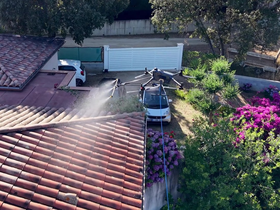 Drone de pulvérisation en intervention au-dessus d'une maison, relié à la cuve par son tuyau d'alimentation